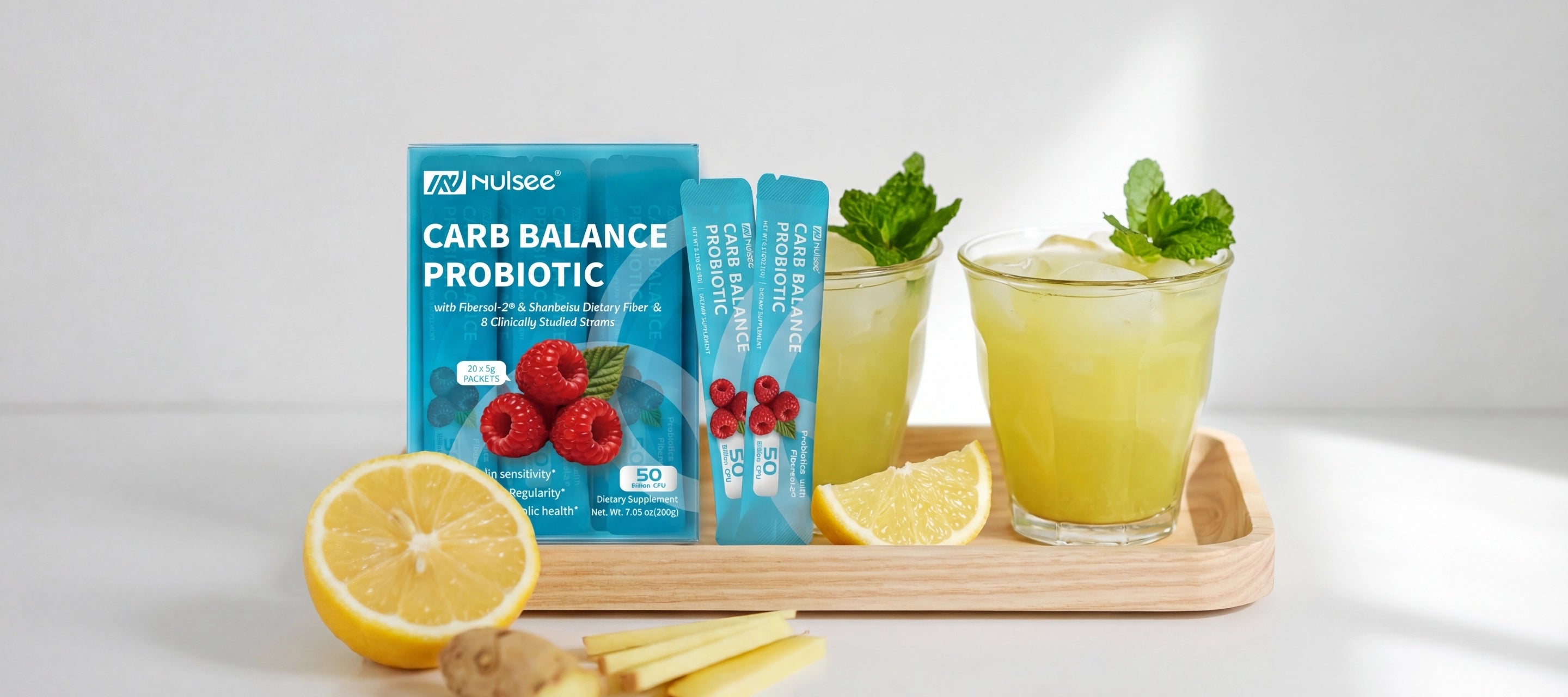 Nulsee Carb Balance Probiotic turquoise box and packets on wooden tray with two glasses of yellow-green healthy drinks garnished with mint leaves, fresh lemon slices and ginger root on light grey background